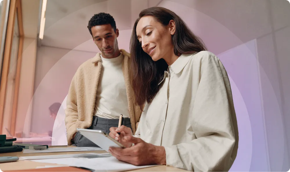 Two people in a modern office setting, one writing on a tablet, the other observing. Both are casually dressed and engaged in discussion.