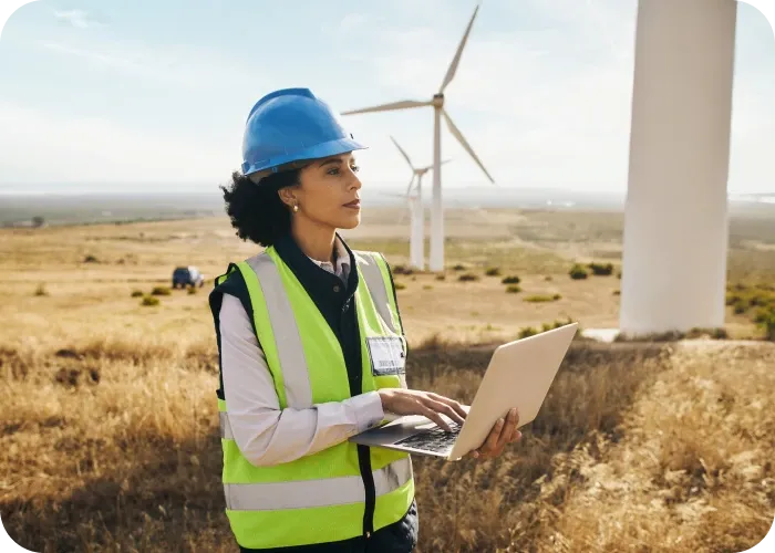Woman in blue hard hat and high-visibility vest using a laptop at a wind farm with turbines on a dry grassy plain.