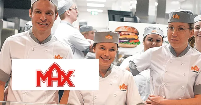 Employees in white uniforms and gray caps stand in a fast-food kitchen with a "Max" logo visible on their attire.
