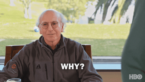 A gif of comedian Larry David. He wears round glasses and a dark grey jacket. He is outside near a golf course, seated down at a table. He shrugs and says