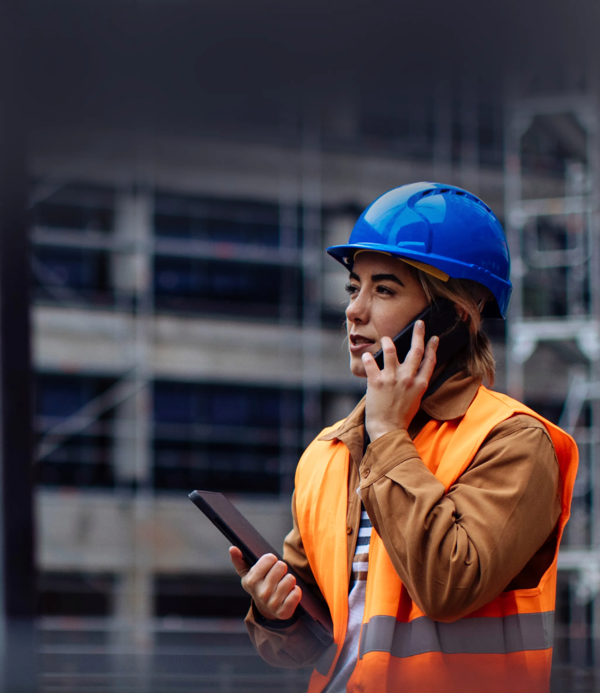 A construction worker in a yellow hard hat and safety vest holds a tablet, looking upwards with buildings blurred in the background.