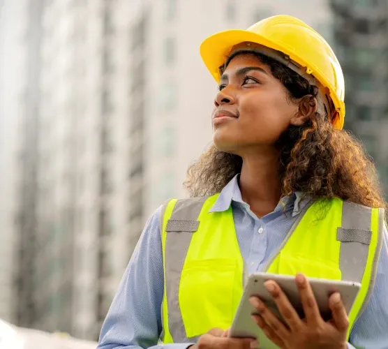A construction worker in a yellow hard hat and safety vest holds a tablet, looking upwards with buildings blurred in the background.