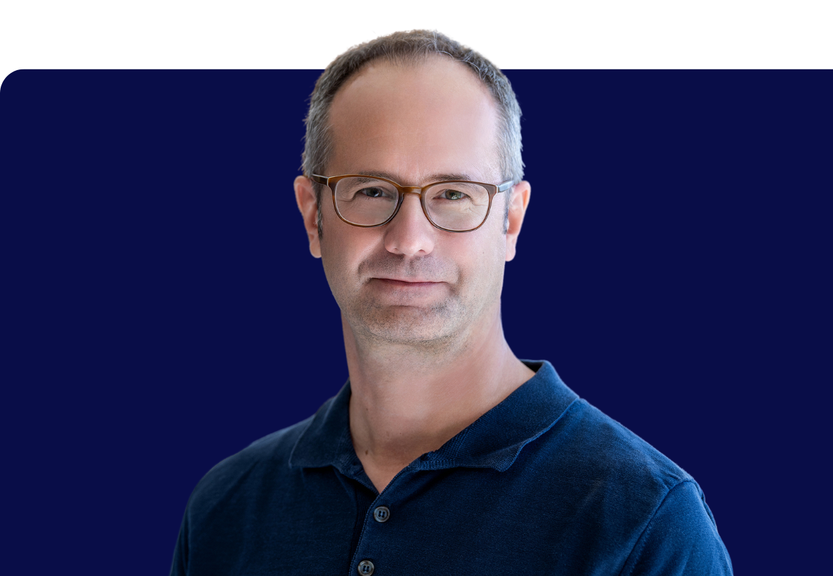 Man with glasses and short hair wearing a dark blue polo shirt against a solid dark blue background.