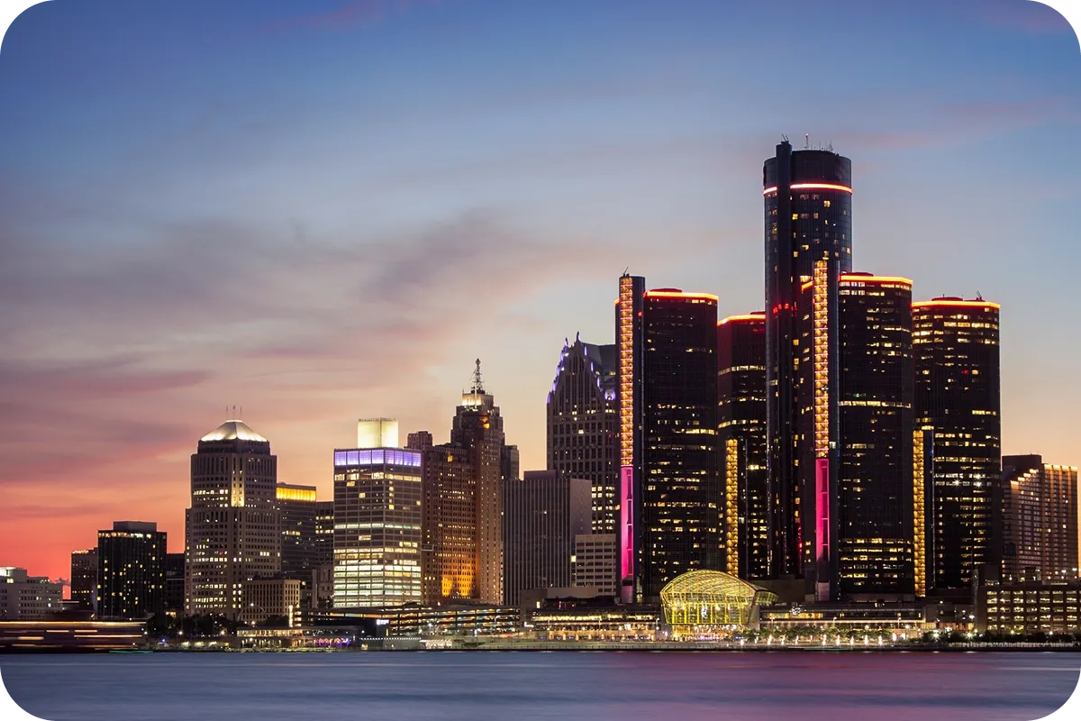 Skyline of a city at dusk with illuminated skyscrapers reflecting in a calm river, under a vibrant, colorful sky.