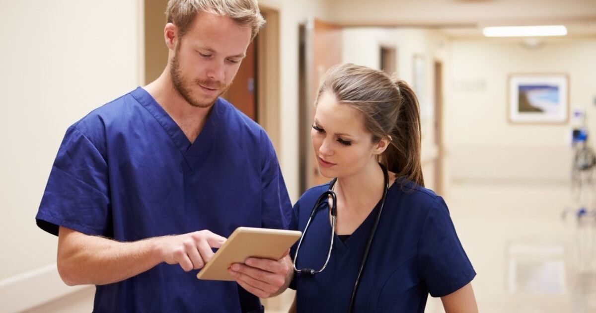 Two healthcare professionals in blue scrubs discuss information on a tablet in a hospital corridor.