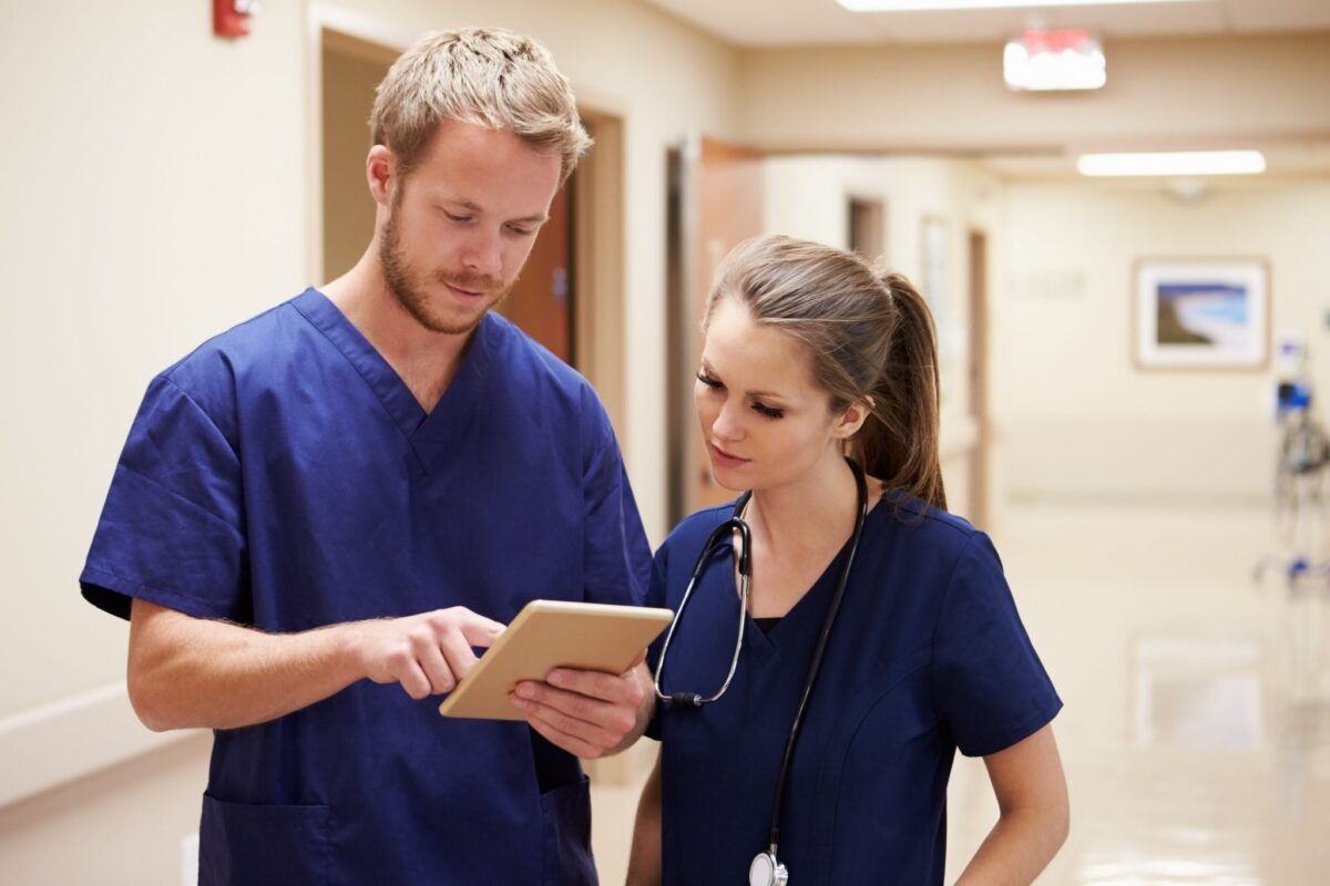 Two healthcare professionals in blue scrubs discuss information on a tablet in a hospital corridor.