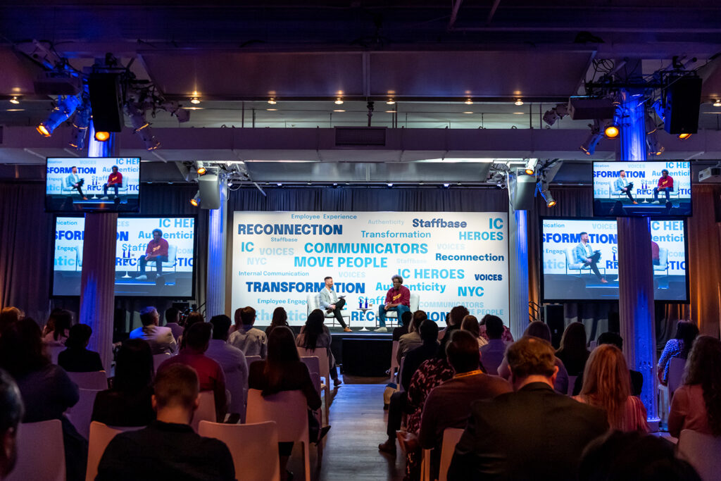 W. Kamau Bell sits on stage with host Adam Brayford at VOICES 2022 in New York City. He looks out into a packed audience and there are words in blue text highlighted behind him that say things like