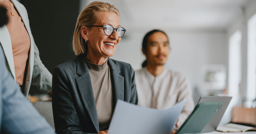 woman in glasses and blazer holding paper and smiling with coworkers surrounding her