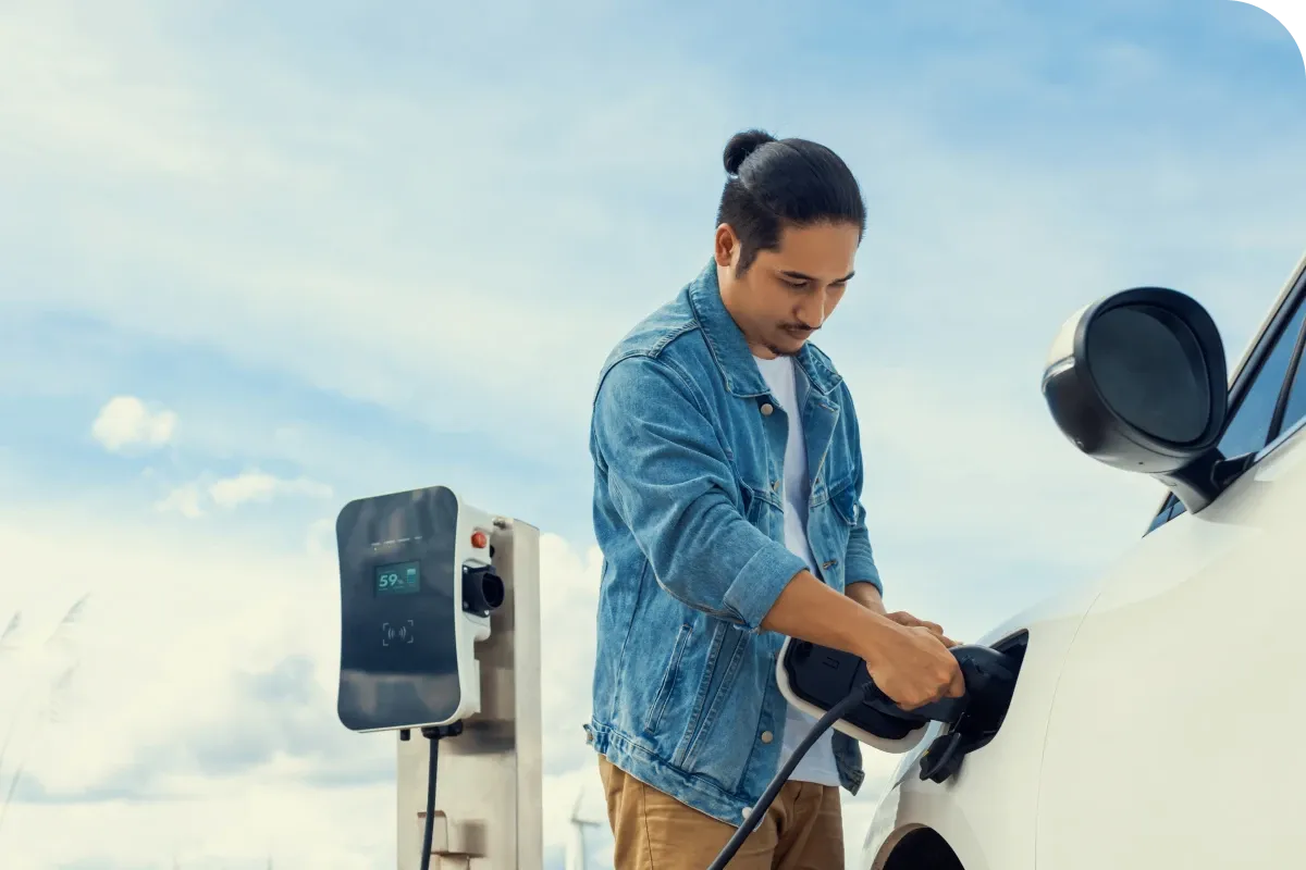 A man in a denim jacket charges an electric car at a charging station under a clear blue sky.