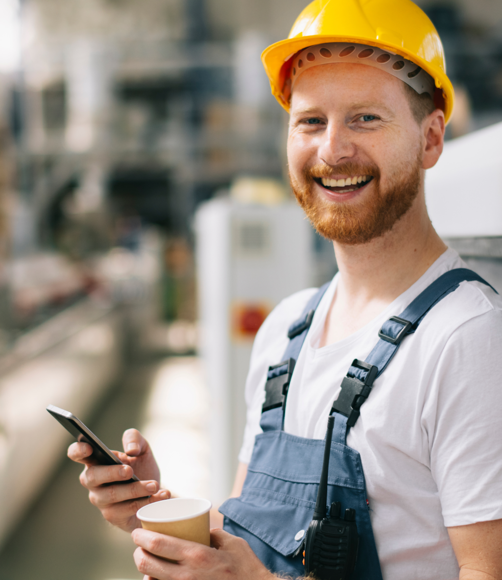 Smiling construction worker in a yellow hard hat, holding a smartphone and a cup, stands in an industrial setting.