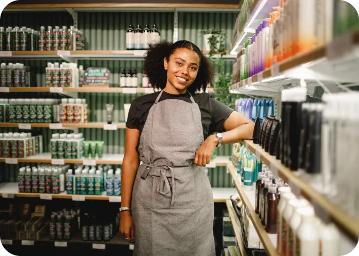 Smiling young woman in gray apron standing in a store aisle, leaning on a shelf stocked with bottles and personal-care products.