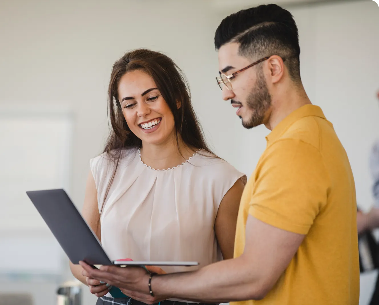 Two people smiling and looking at a laptop together in a bright room. One is holding the laptop, and they appear to be discussing something.