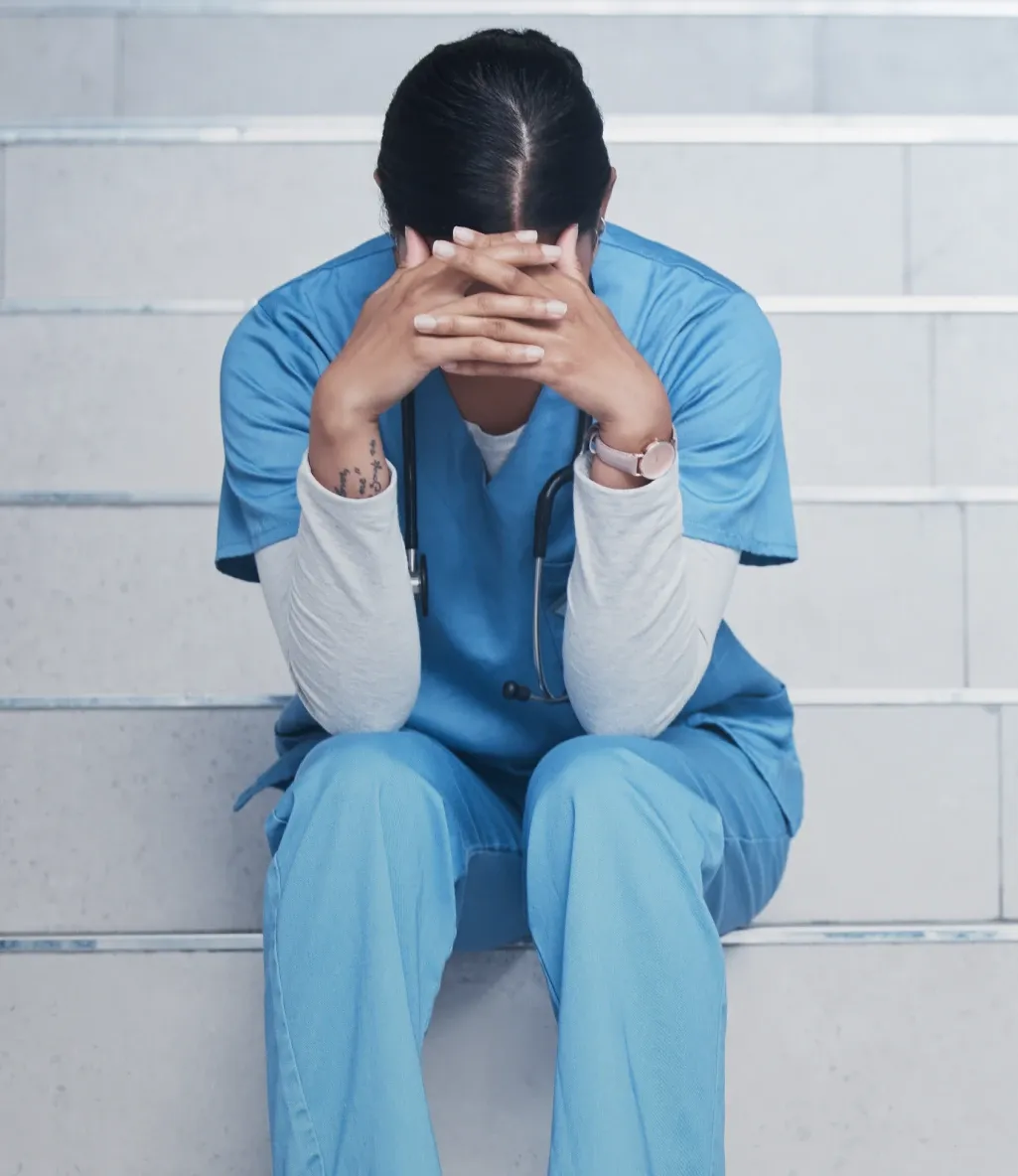 A person in blue scrubs sits on stairs, head in hands, appearing stressed. A stethoscope hangs around their neck.