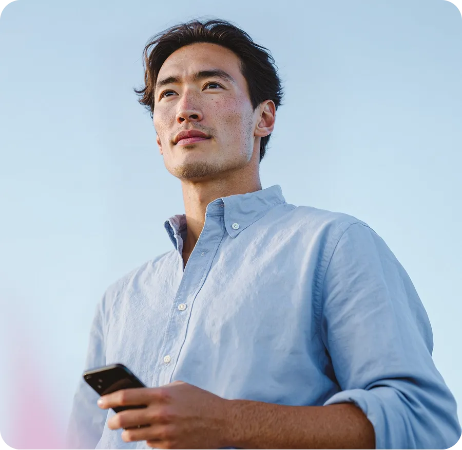 A person in a light blue shirt holds a smartphone, looking thoughtfully into the distance against a clear blue sky.