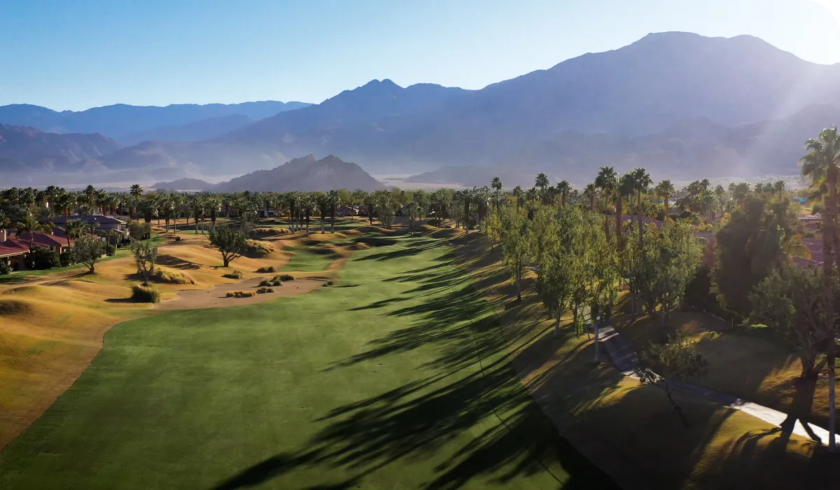 Aerial view of a lush golf course with palm trees, surrounded by mountains under a clear blue sky.