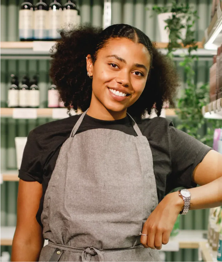 Smiling woman in a gray apron stands in a store, surrounded by shelves with bottles and plants in the background.