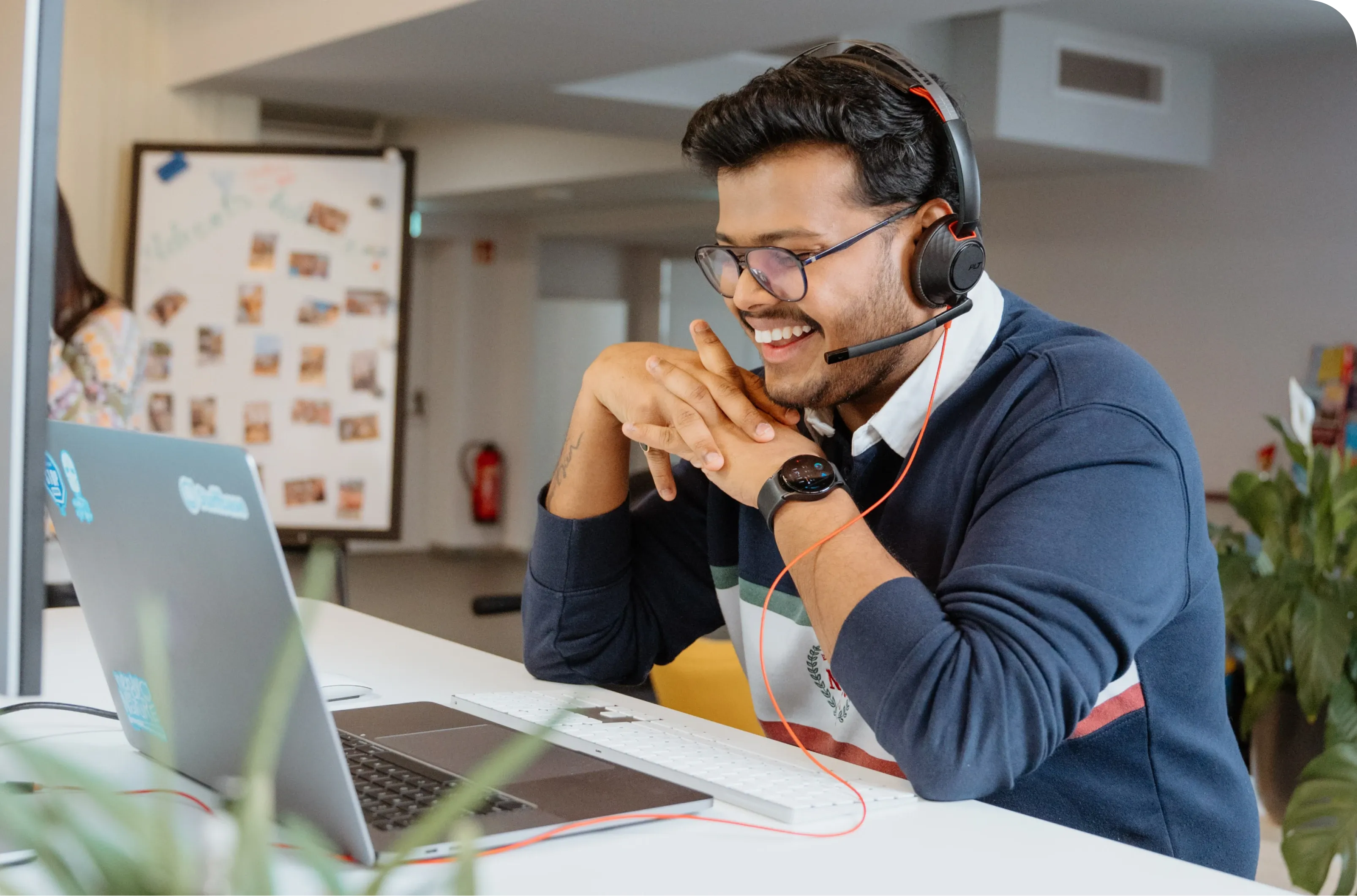Man wearing glasses and a headset, smiling at a laptop in a bright office space with plants and a whiteboard in the background.