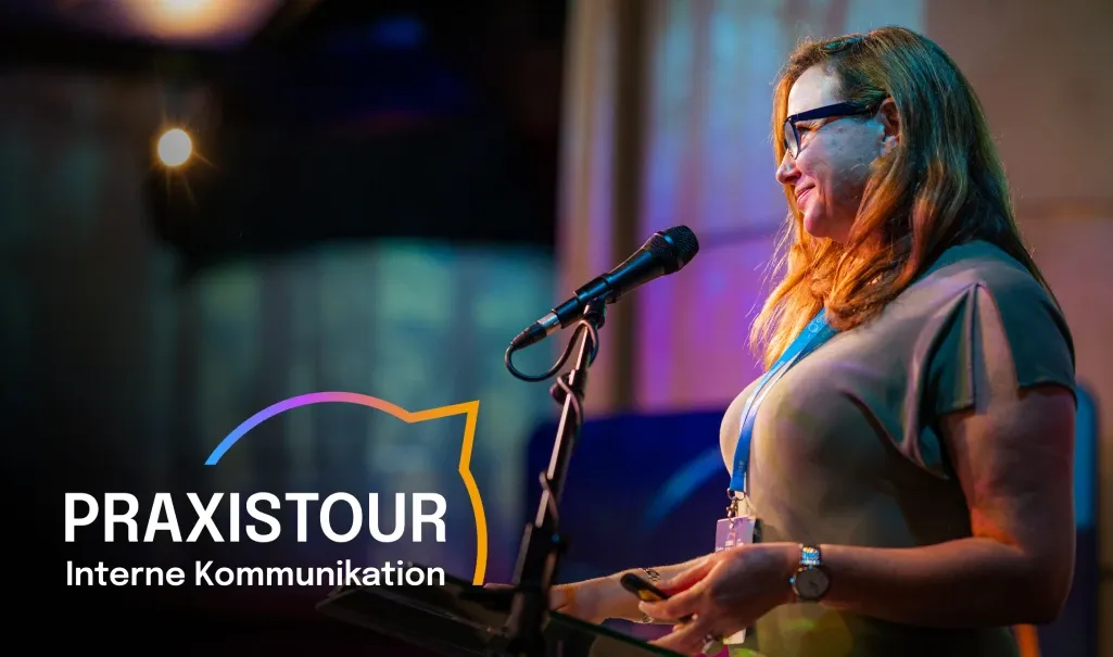 Woman speaking at a podium with a microphone, wearing glasses. "COMMS CLUB" text with colorful outline is visible on the left.