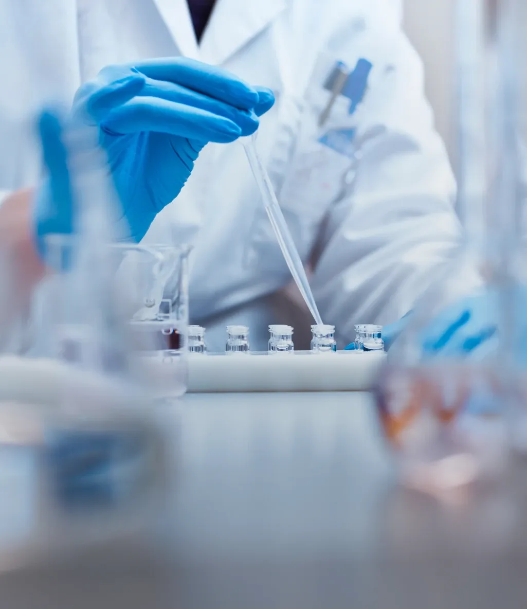 Scientist in lab coat and blue gloves using a pipette to transfer liquid into small vials in a laboratory setting.