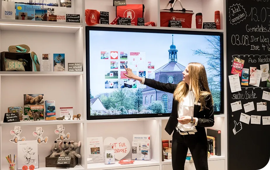 A woman in a black blazer presents information on a large screen in a store, surrounded by travel brochures and souvenirs.
