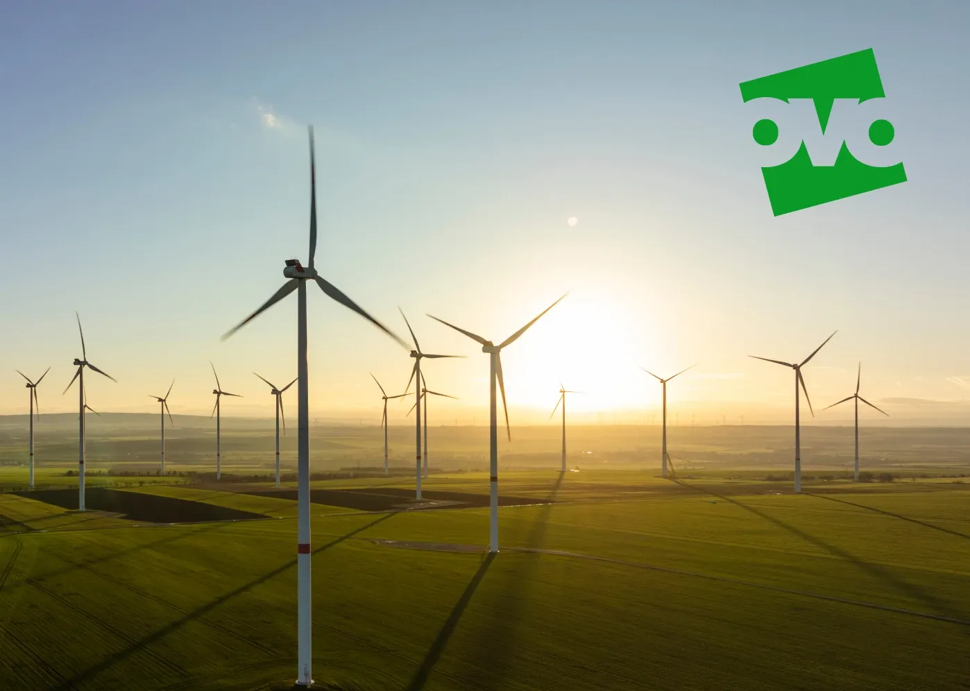 Wind turbines on a grassy field at sunset, with the sun low on the horizon and a green logo in the sky.
