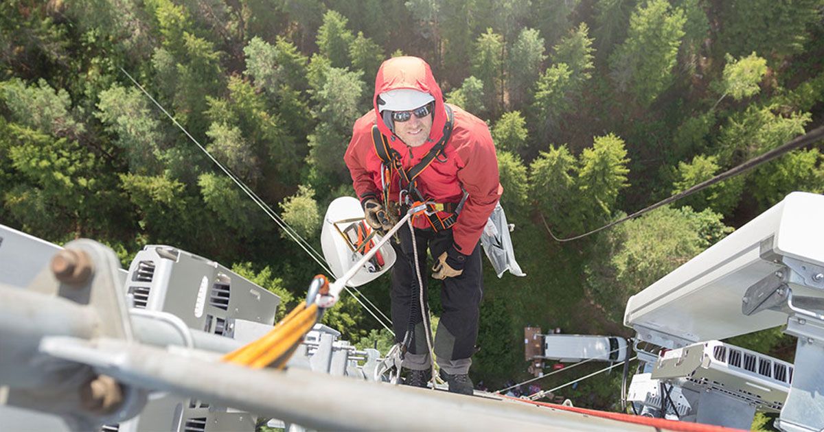 A worker in a red jacket and helmet climbs a tall communication tower, surrounded by forested landscape below.