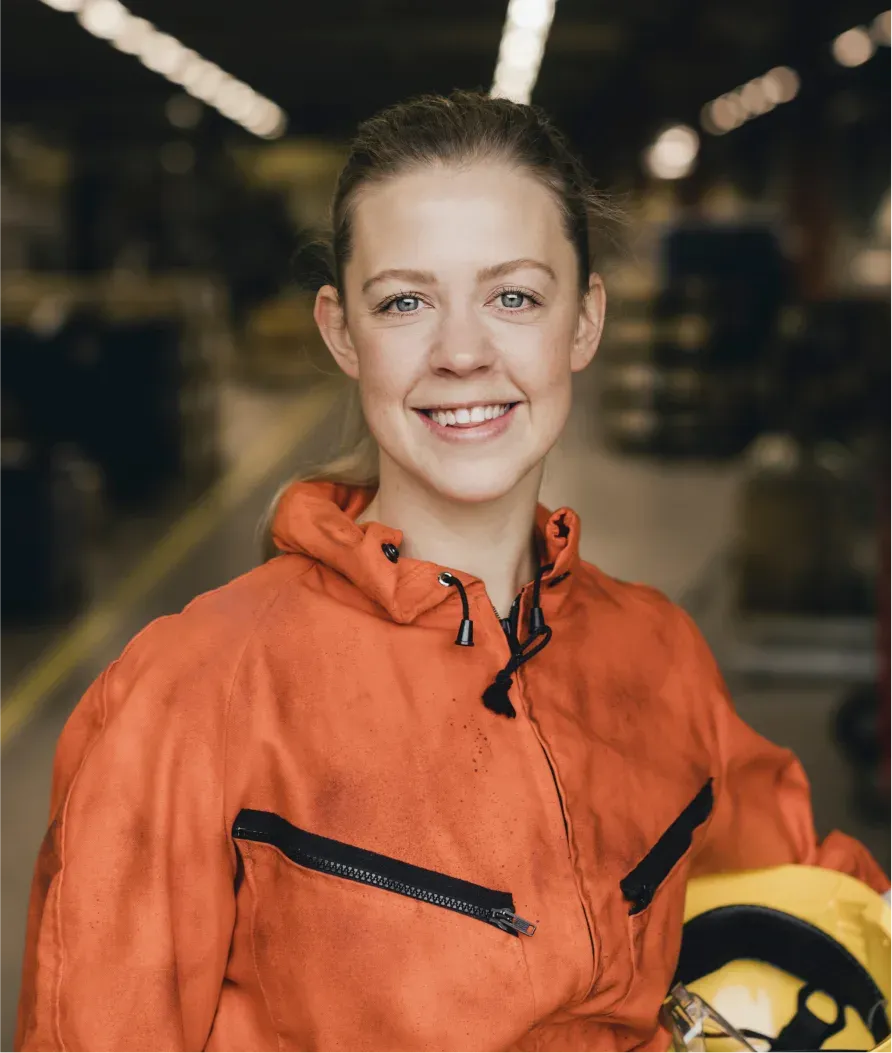 Smiling person in an orange work jumpsuit holds a yellow hard hat, standing in an industrial setting with blurred background.