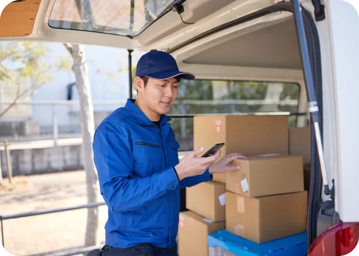Delivery worker in blue uniform checks his phone at an open van loaded with stacked cardboard boxes.