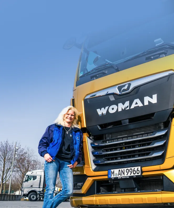 Person in a blue jacket and jeans stands smiling next to a large yellow truck labeled "WOMAN" with a clear blue sky above.