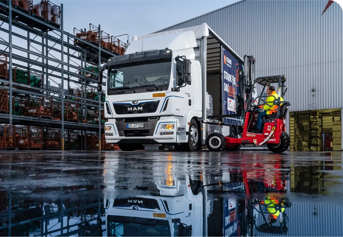 A white truck parked in an industrial area with a forklift unloading cargo beside it, reflecting on a wet surface.