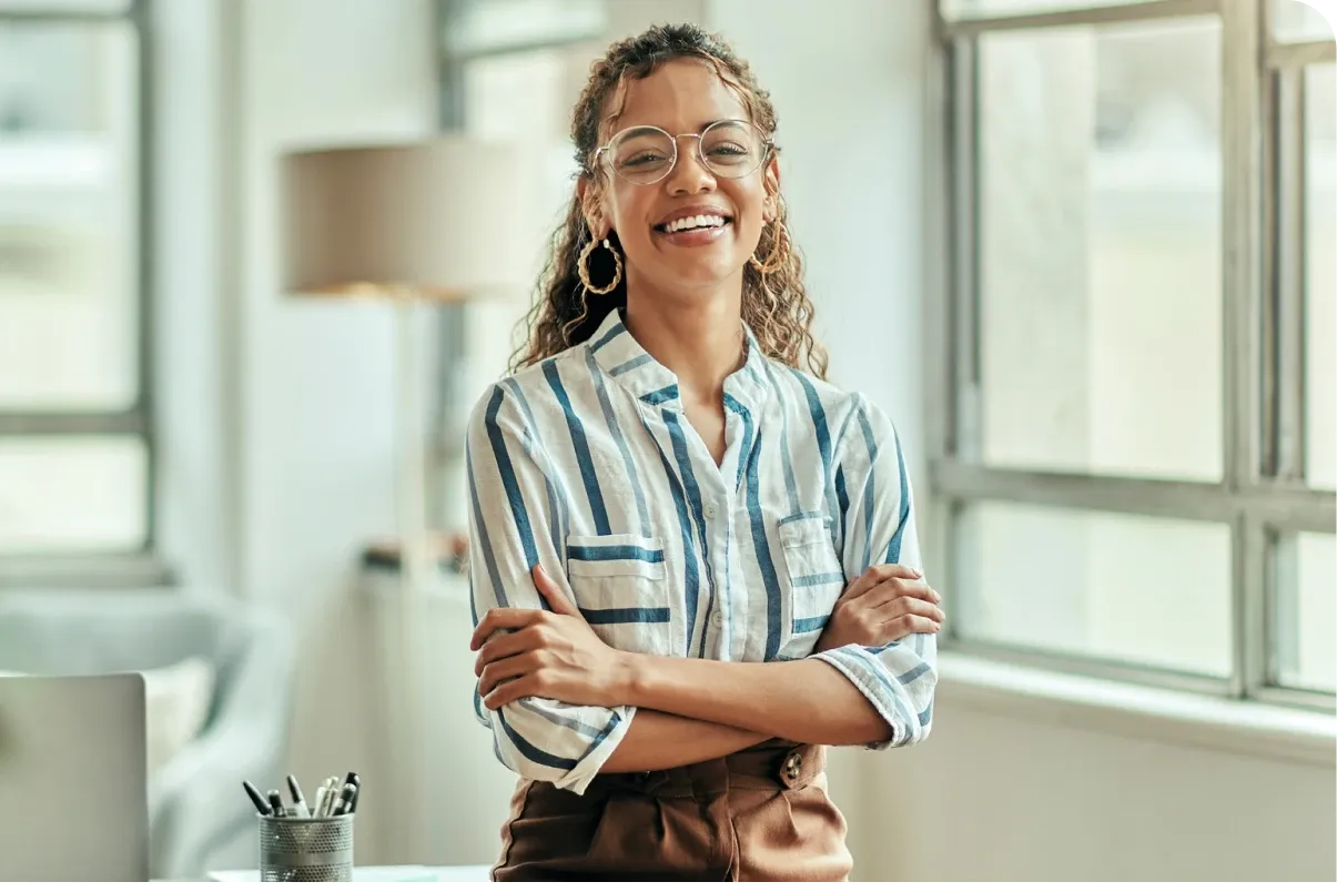 Smiling person with glasses and curly hair, wearing a striped shirt, stands with arms crossed in a bright room with large windows.