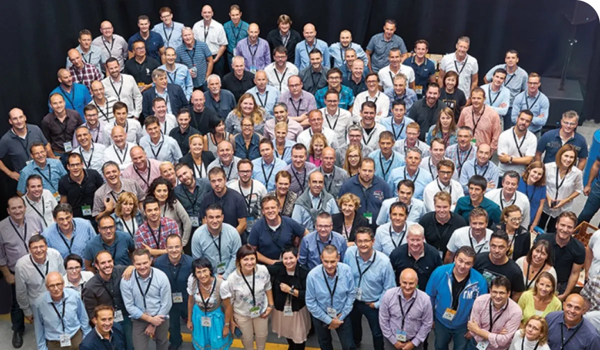 A large group of people, wearing name tags, stand closely together, smiling at the camera in a well-lit indoor setting.