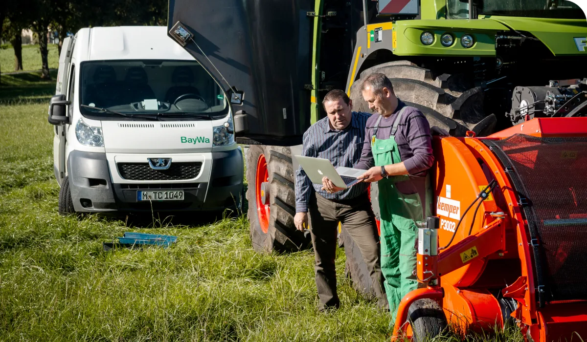 Two men stand in a grassy field, examining a laptop. Nearby are a white van and large agricultural machinery.