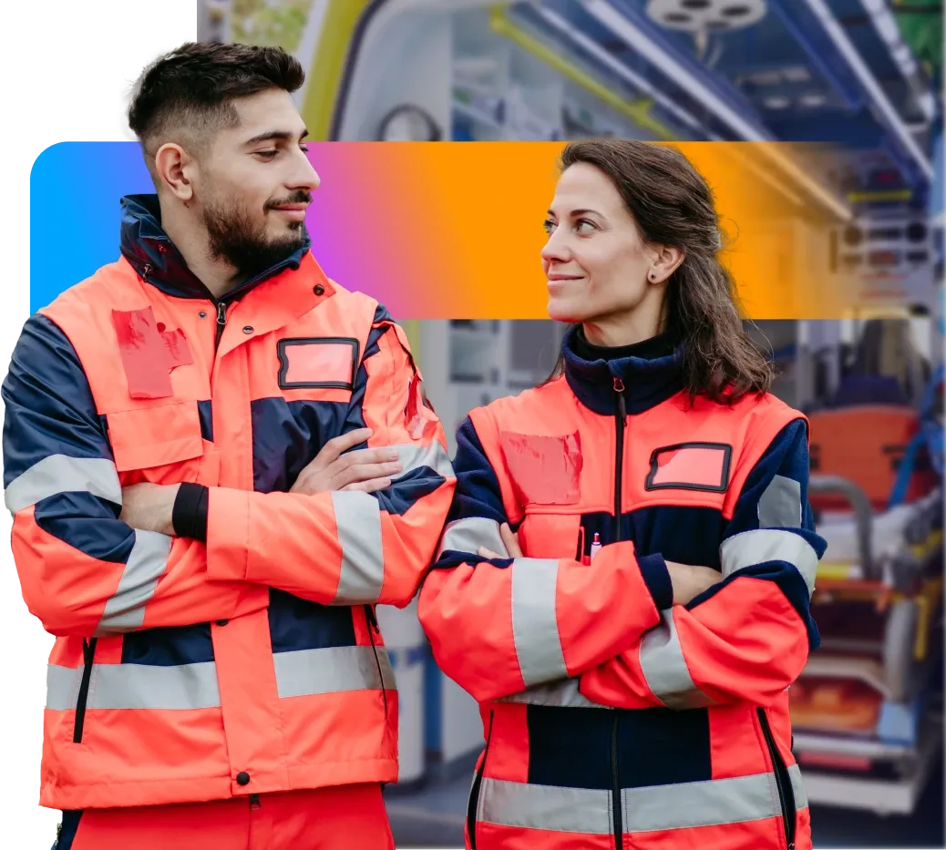 Two people in bright orange and navy uniforms stand with arms crossed, smiling at each other, in front of an ambulance interior.
