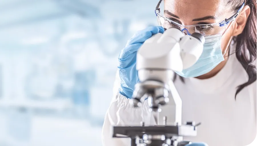 A scientist wearing safety goggles and a mask examines a sample through a microscope in a laboratory setting.