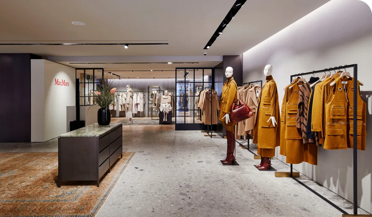 Fashion store interior of Max Mara with mannequins in camel-colored coats, a display table, and racks of clothing. Modern design with soft lighting.