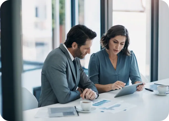 Two colleagues reviewing a tablet and charts at a bright office table with coffee cups.