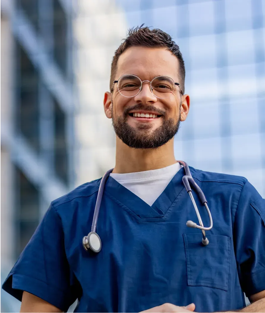 Smiling healthcare professional in blue scrubs and glasses, with a stethoscope around his neck, standing in front of a glass building.