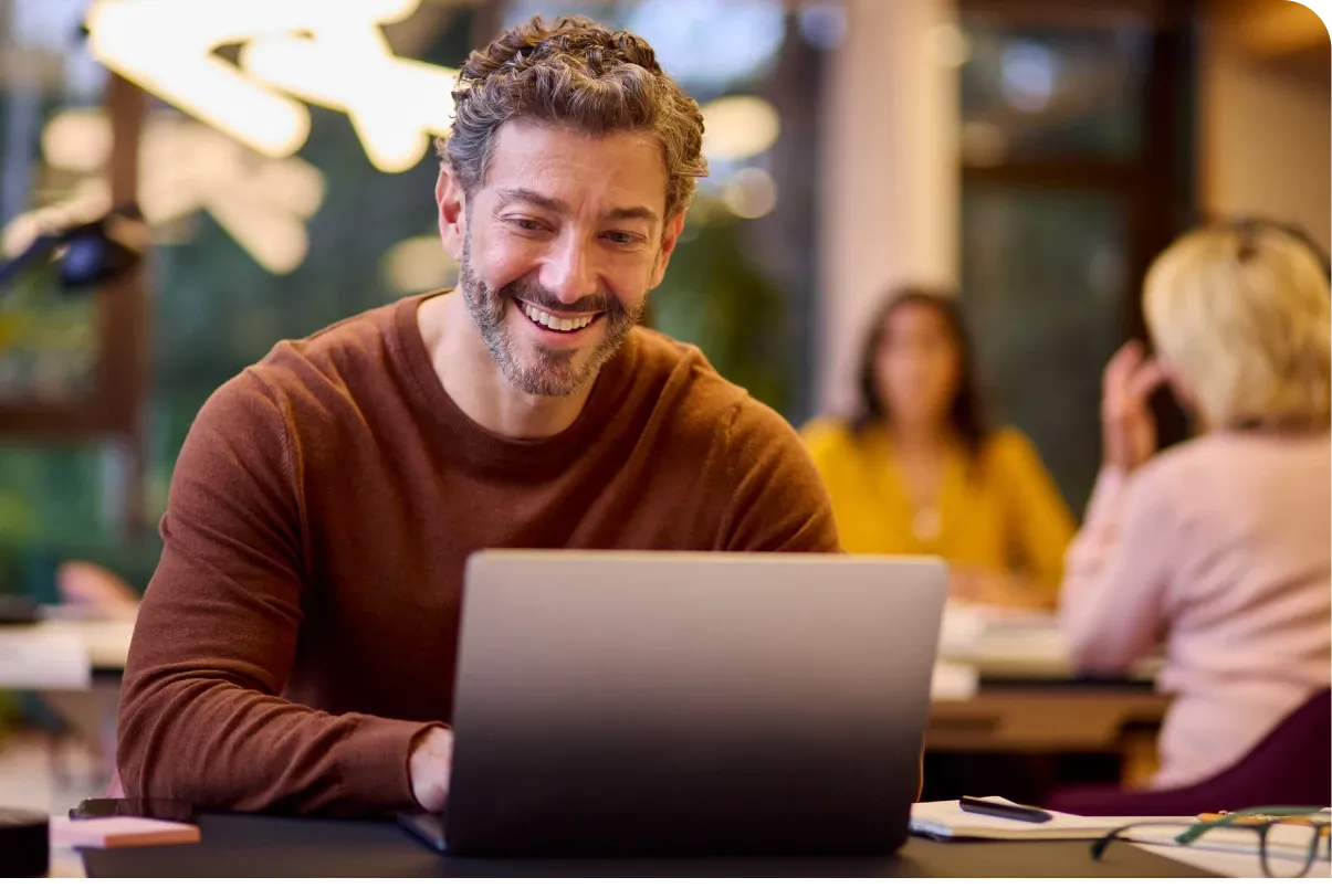 A man with a beard smiles at a laptop in a cozy office. Two people are blurred in the background.