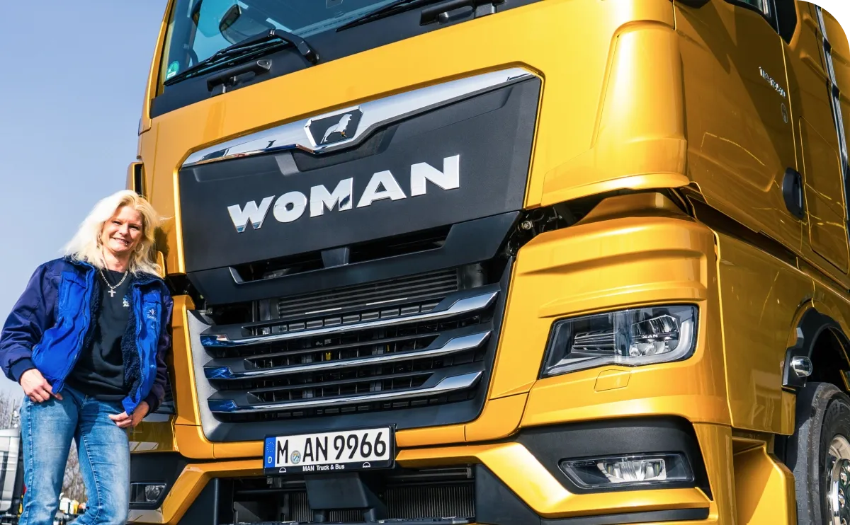 Person smiling next to a large yellow truck with "WOMAN" on the front. The truck has a visible license plate "M AN 9966".