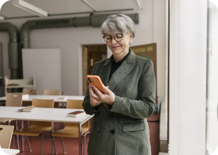 Smiling older woman in green blazer and glasses standing in a classroom, looking at an orange smartphone.