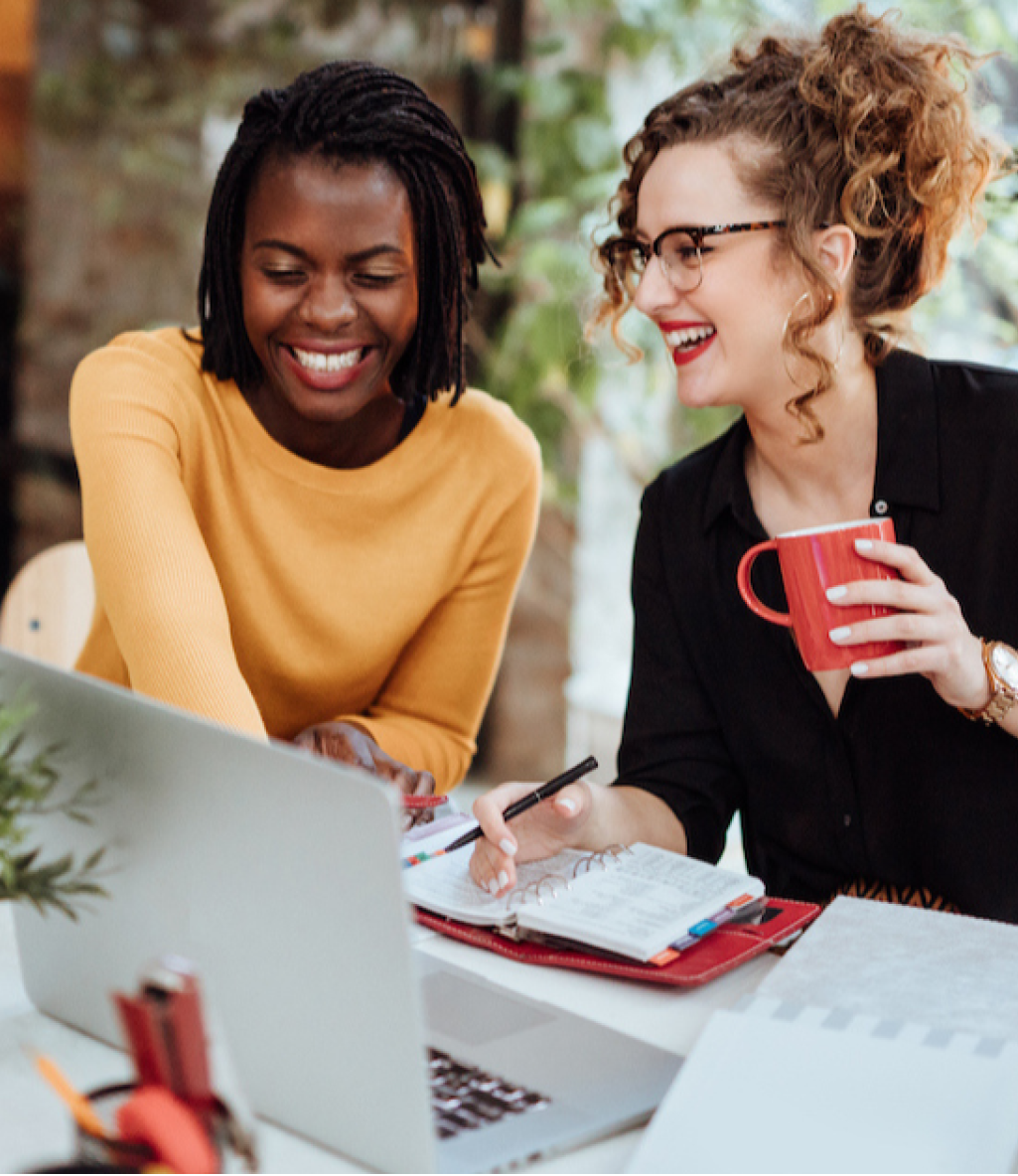 Two women smiling and working together at a desk with a laptop, notebook, and a red mug, surrounded by plants.