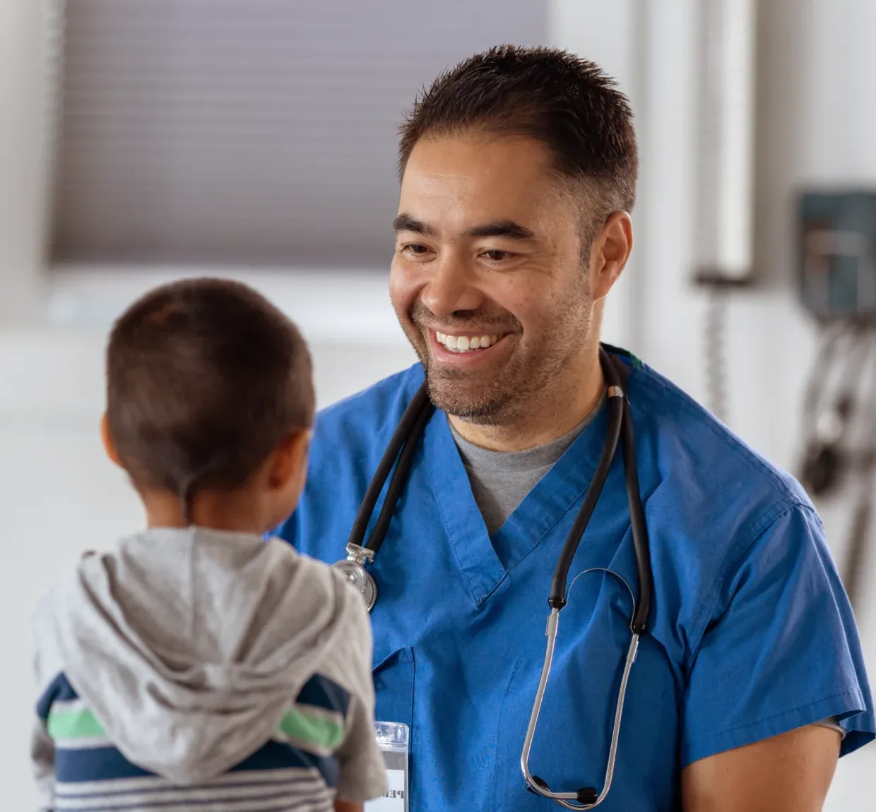 A smiling healthcare professional in blue scrubs interacts with a young child in a medical setting.