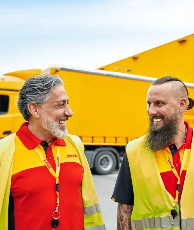 Two DHL workers in branded uniforms and safety vests smile at each other, standing in front of a yellow delivery truck.