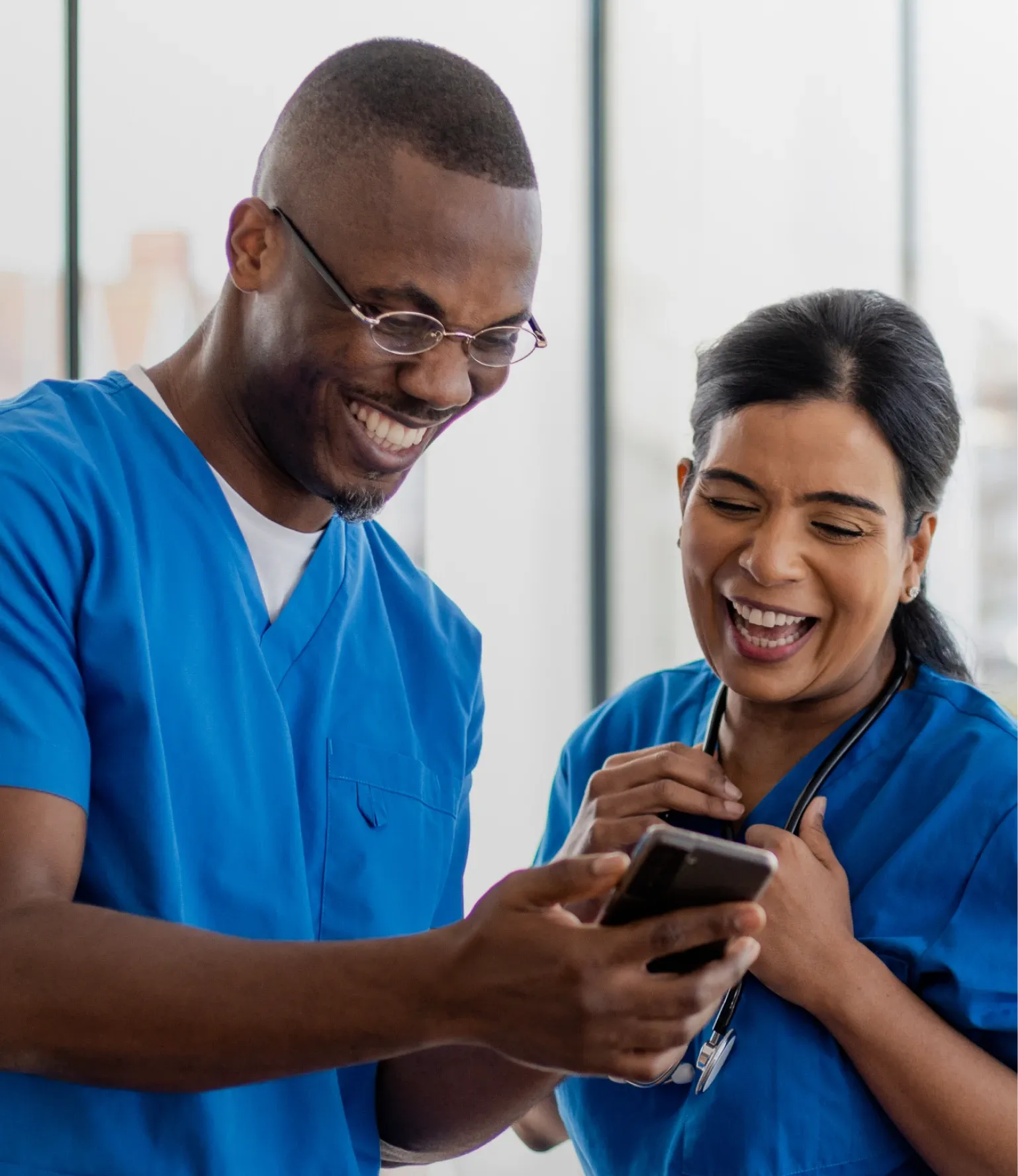 Two healthcare professionals in blue scrubs smiling and looking at a smartphone together, standing in a brightly lit room.