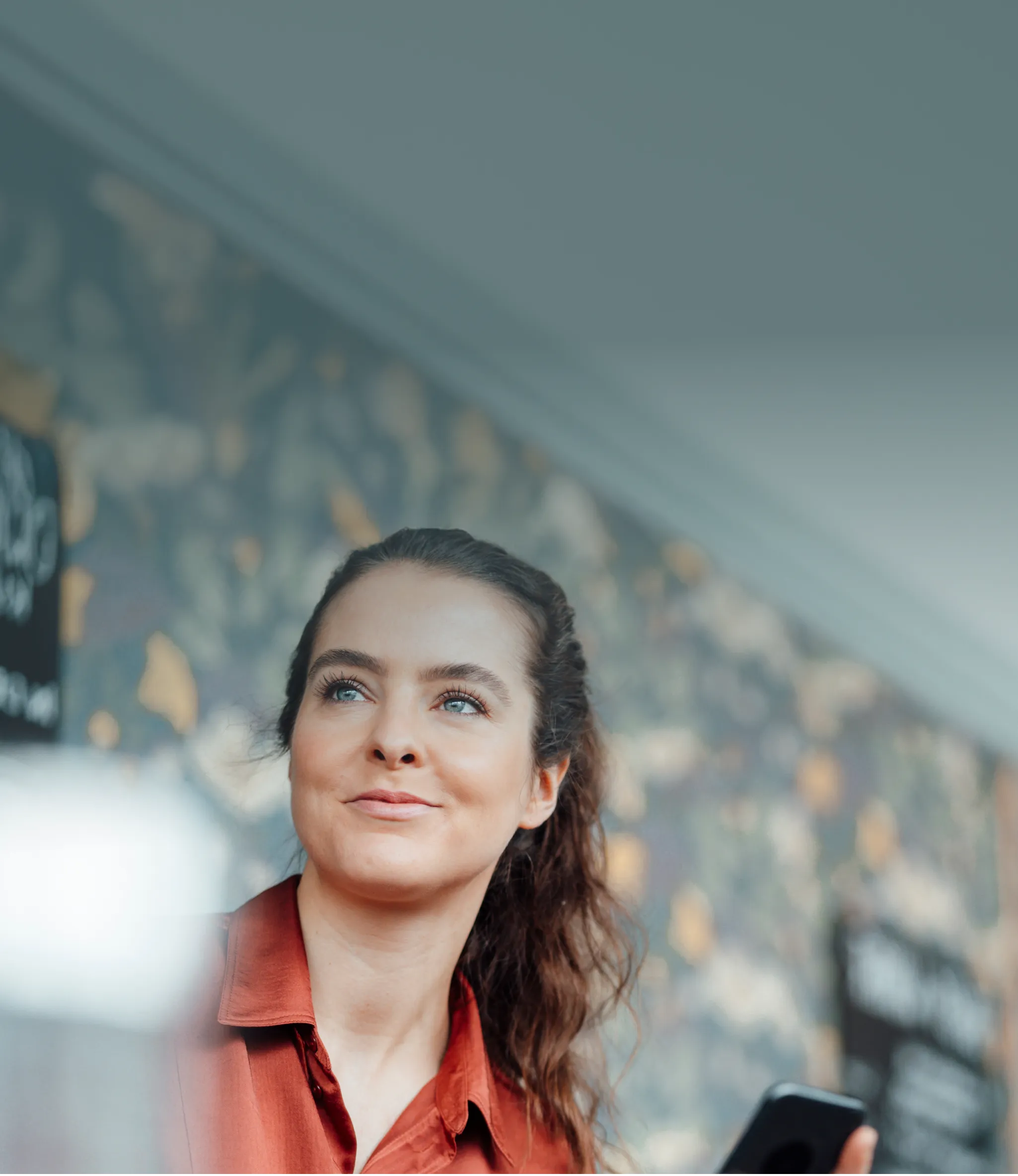 Woman with a ponytail in a red shirt holding a phone, looking to the side, with a floral-patterned wall in the background.