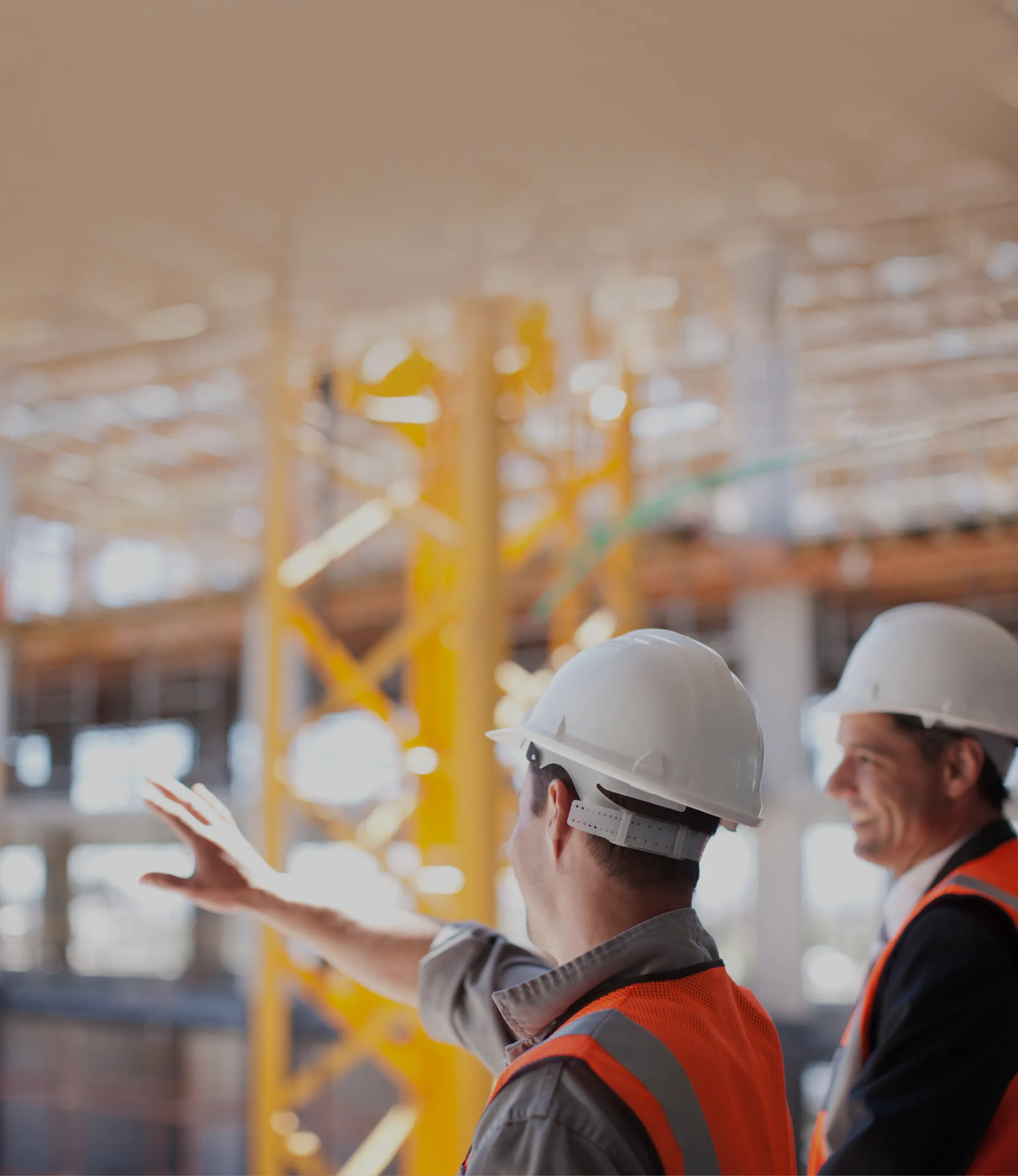Two construction workers in safety gear and helmets discuss a project on-site, with scaffolding and equipment in the background.