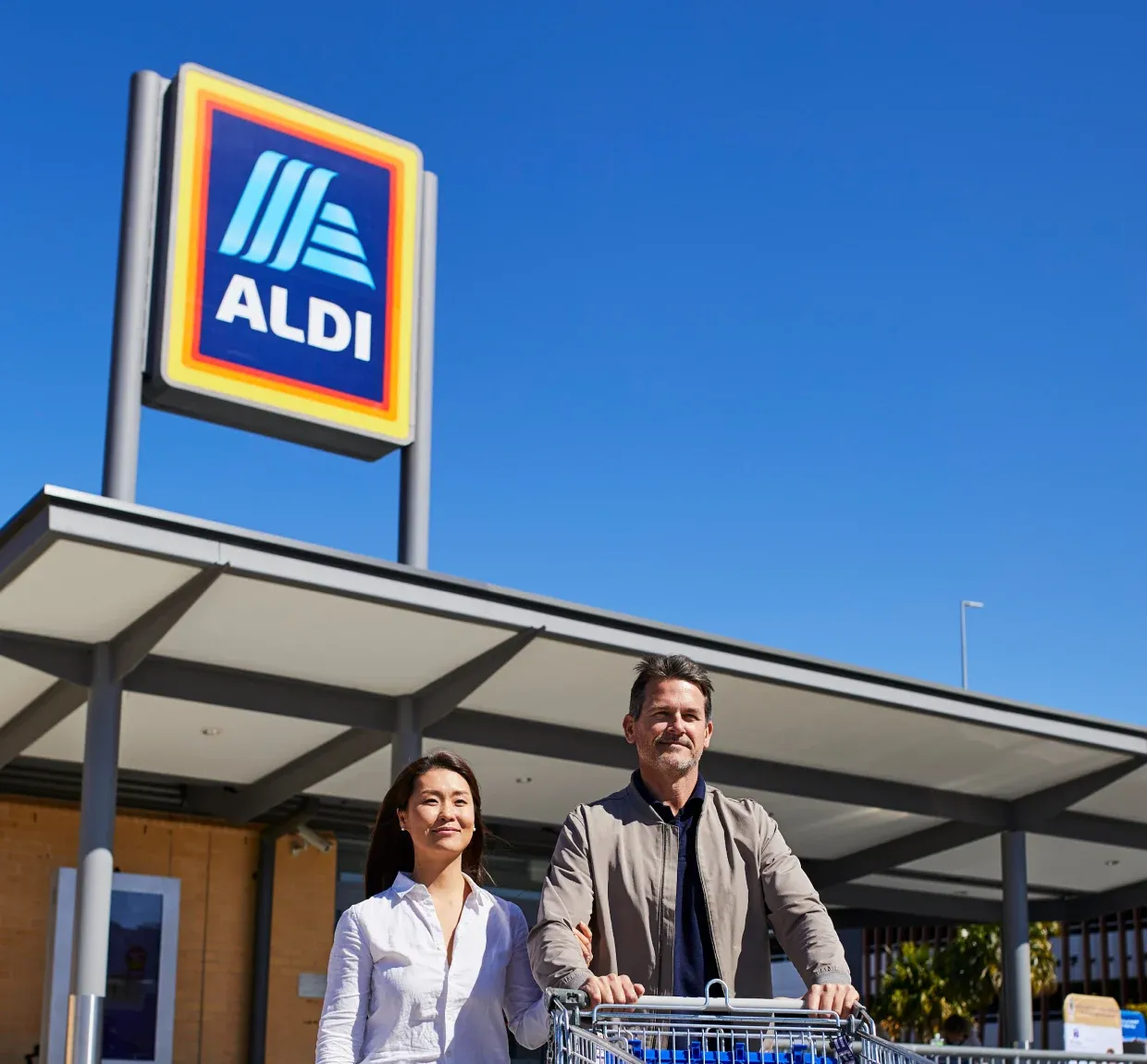 A man and woman stand with a shopping cart in front of an Aldi store under a clear blue sky.