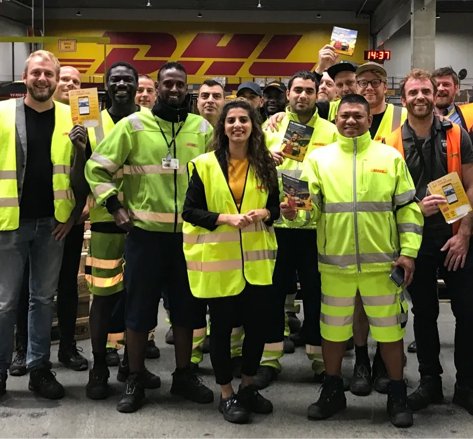 A diverse group of DHL employees in high-visibility uniforms pose together, smiling, in a warehouse setting.