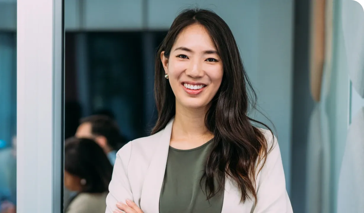 Smiling woman with long hair in a white blazer and green top, standing confidently with arms crossed in an office setting.